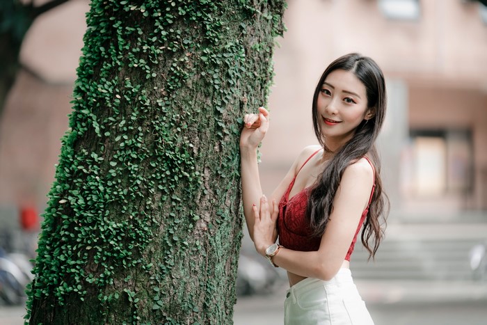 Asian woman in red top and white shorts by tree trunk
