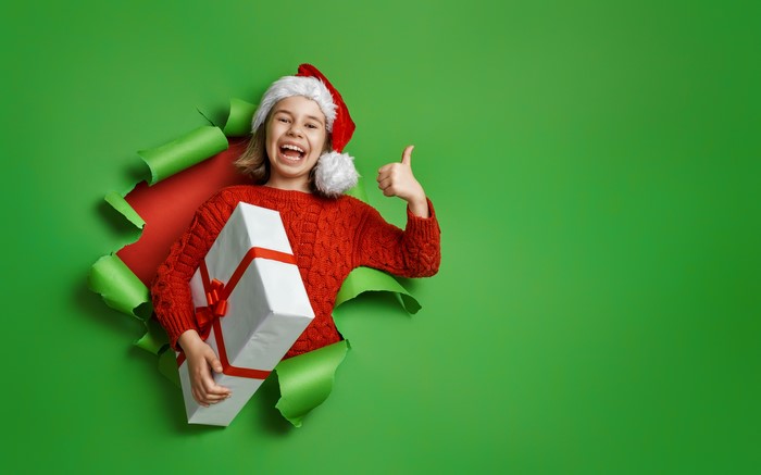 Girl in Santa hat holding Christmas gift, giving thumbs up