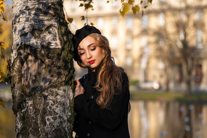 Woman in black coat and beret leaning on birch tree