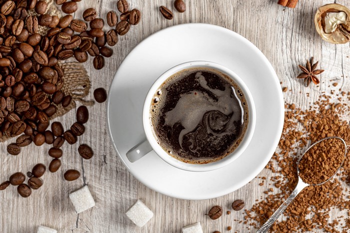 Coffee beans, cup of coffee, sugar cubes, and spices on wood