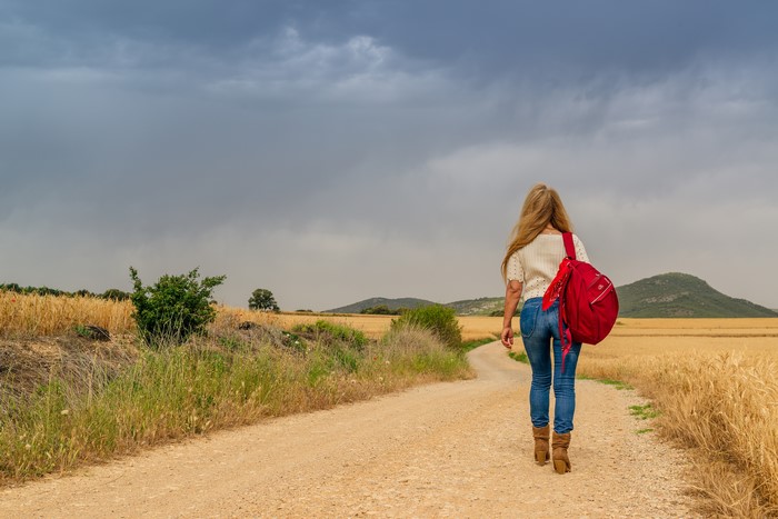 Woman with red backpack walking on dirt road through fields