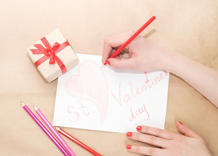 Hands writing "Valentine's Day" on paper with gift and pencils