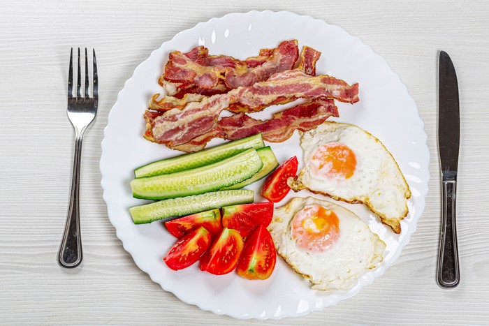 Fried eggs with bacon, cucumbers, and tomatoes on a plate with fork and knife