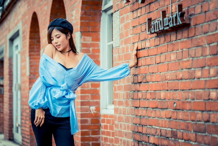 Asian woman in blue blouse and beret leaning on brick wall