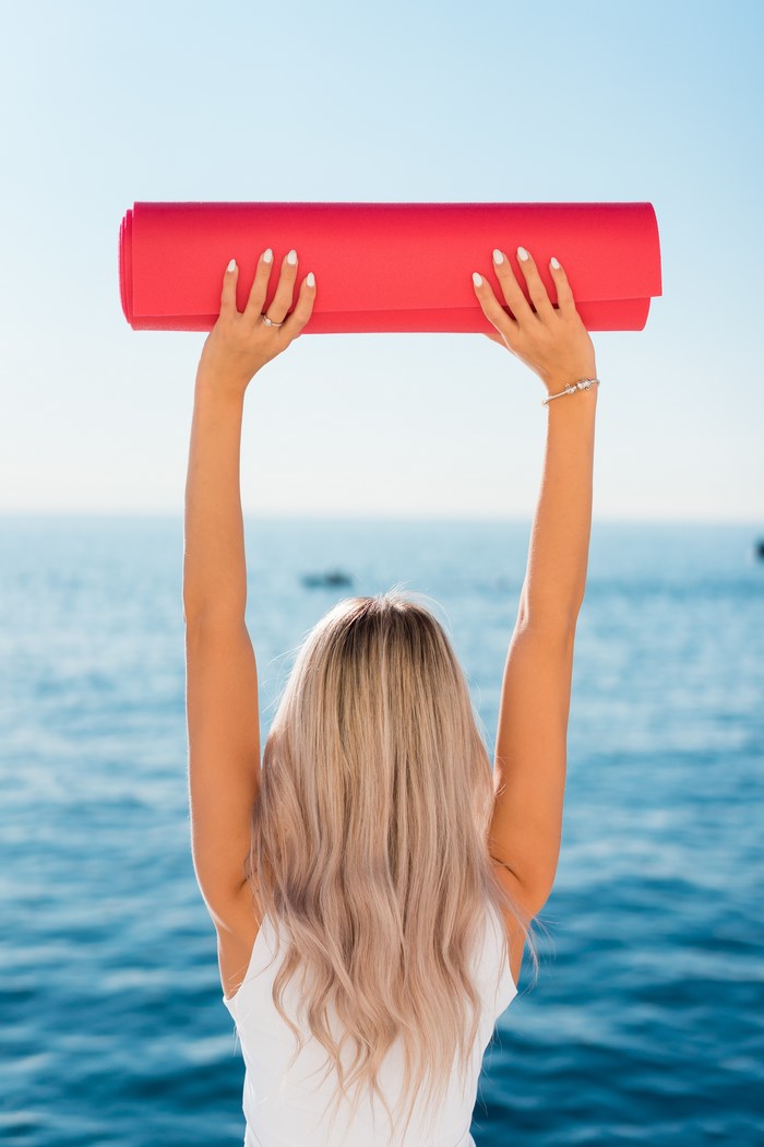 Blonde woman holding a red yoga mat over the ocean