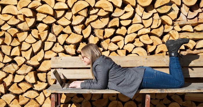 Woman with laptop on bench in front of woodpile