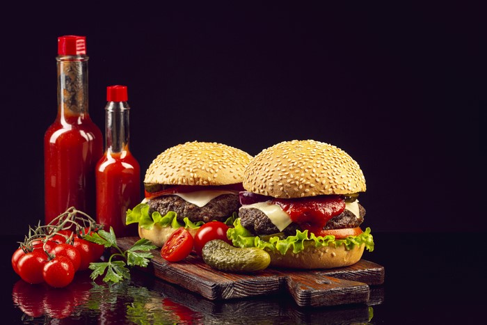 Two hamburgers with ketchup bottles and tomatoes on a cutting board