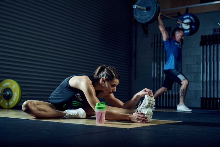 Woman stretching in gym with man lifting barbell