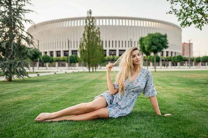 Blonde girl in a dress sitting on grass in front of a stadium