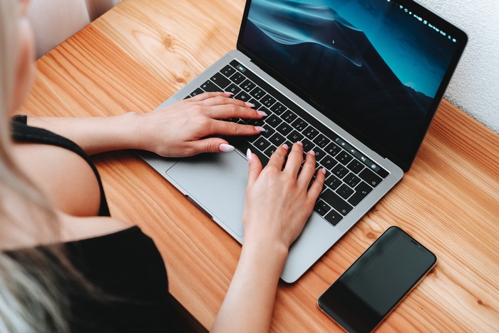 Woman typing on a laptop with a smartphone