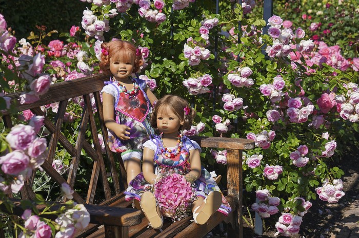 Two dolls sitting on a bench in a rose garden
