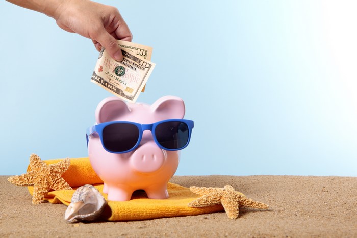 Piggy bank with sunglasses on beach, hand inserting money