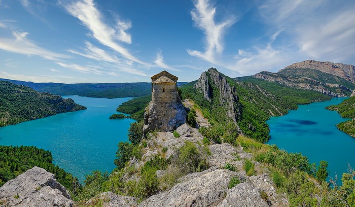 Stone hermitage on a rocky outcrop overlooking a turquoise reservoir in Spain