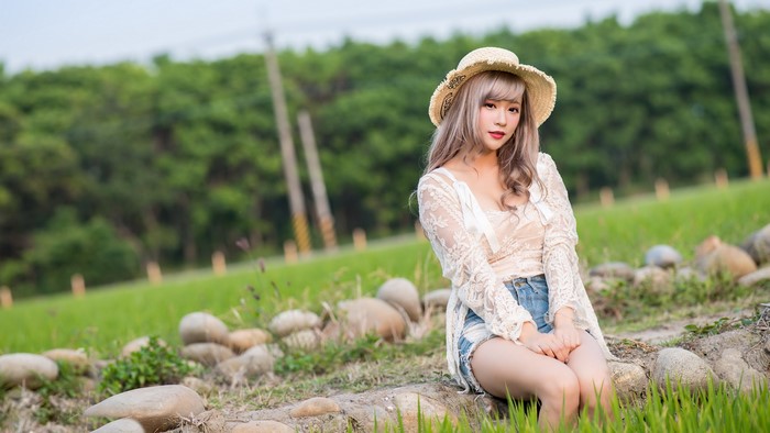 Asian woman with dark blonde hair wearing a hat and white lace top sitting in a field