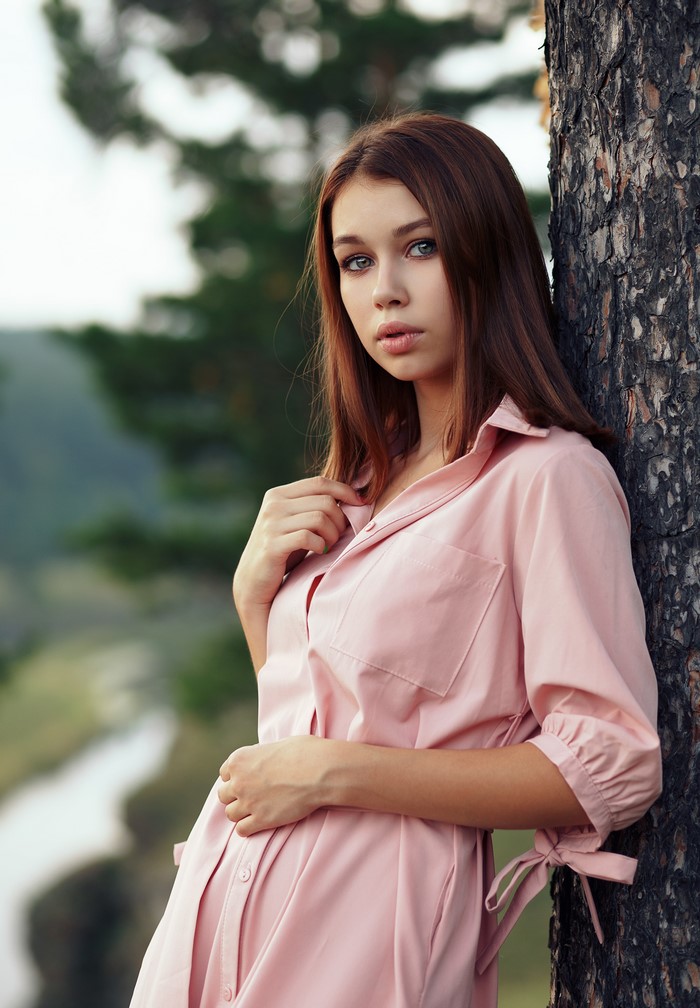 Brown-haired girl in pink dress leaning against a tree