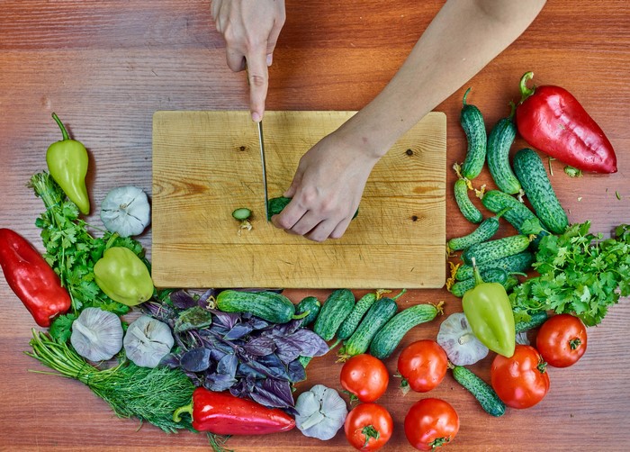 Hands chopping cucumber on cutting board with vegetables