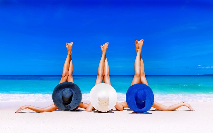 Three women in hats on a beach with legs up