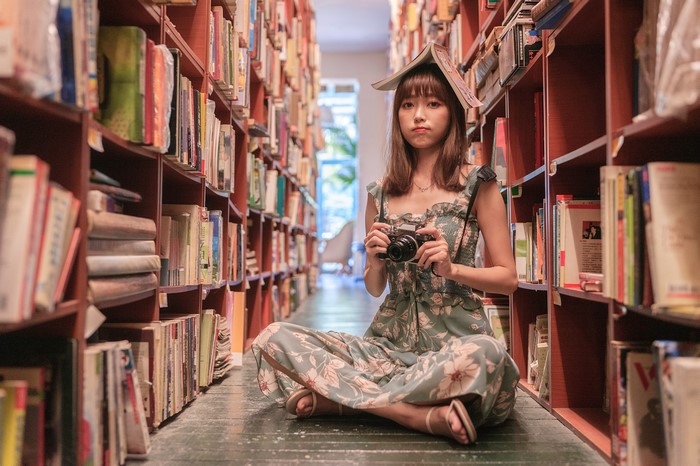 Asian woman with book on head holding camera in library