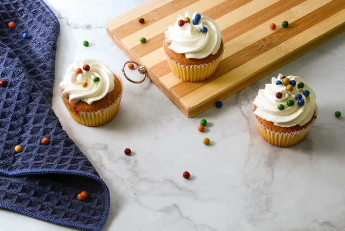 Three cupcakes with white frosting and candy on a marble surface with a cutting board