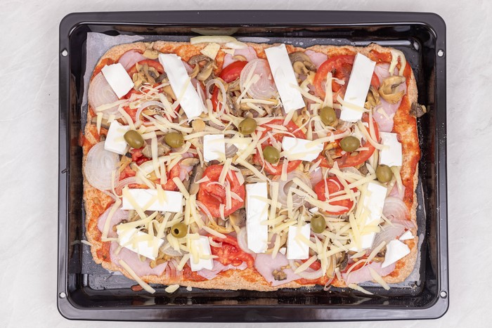 Rectangular pizza with ham, mushrooms, tomatoes, onions, olives, and cheese on a baking sheet