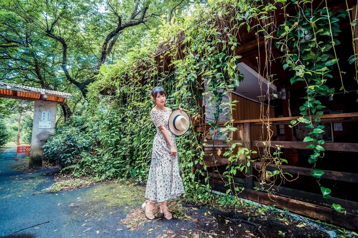 Asian woman in dress holding hat near building covered in vines