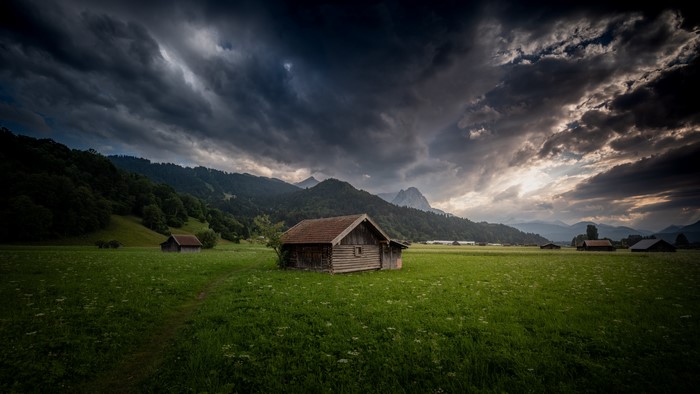Wooden huts in a field with mountains and stormy clouds