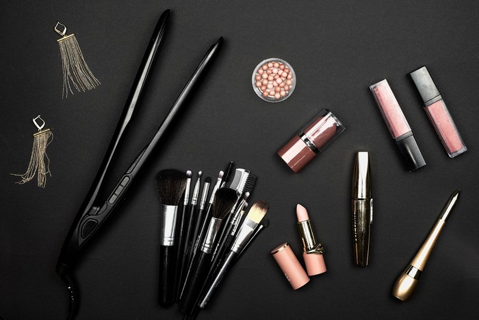 Flat iron, makeup brushes, earrings, and cosmetics on black background