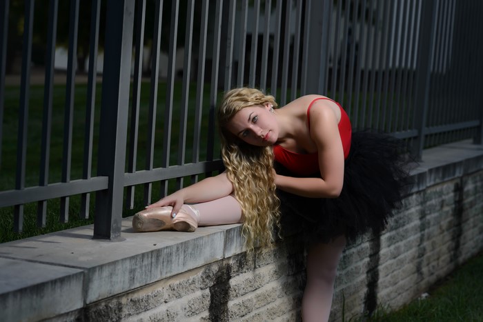 Blonde ballerina in red leotard and black tutu stretching by fence