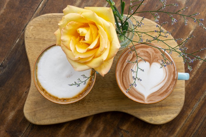 Two coffees with a yellow rose on a wooden board