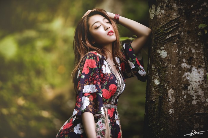 Asian woman in floral dress leaning against tree trunk