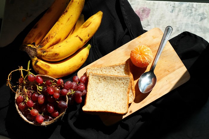 Bananas, Grapes, Bread, Mandarin, and Spoon on Cutting Board