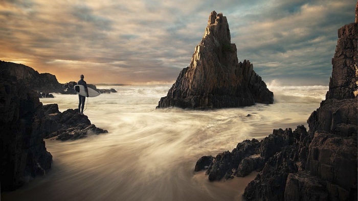 Surfer with surfboard on rocky coast at sunset
