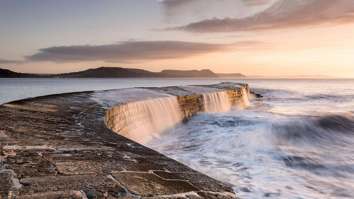Curved stone pier with waves crashing at sunset