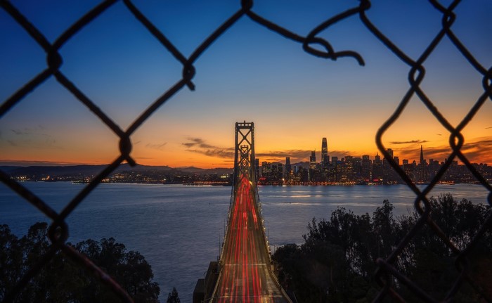 Oakland Bay Bridge and San Francisco cityscape at sunset through a fence