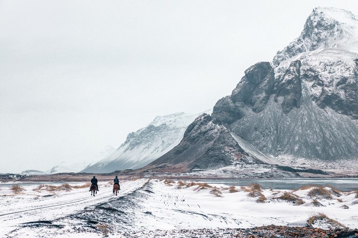 Two people on horseback on a snowy path in Iceland with mountains