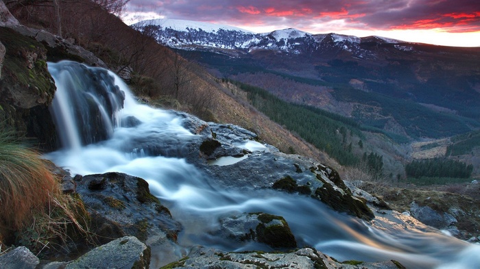 Waterfall cascading down rocks with snow-capped mountains at sunset