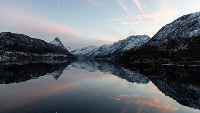 Snow-capped mountains reflected in a fjord under a pastel sky with a crescent moon