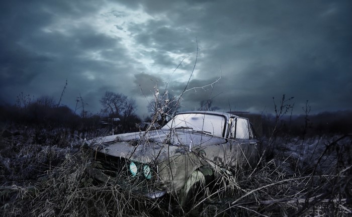 Abandoned car in overgrown field under stormy sky