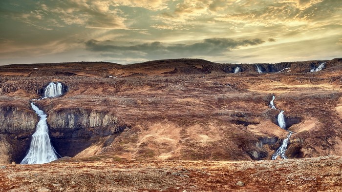 Multiple waterfalls cascade down a rocky, barren hillside under a cloudy sky.