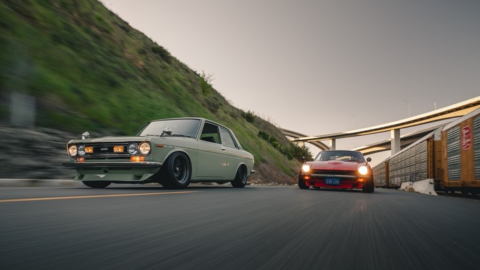 Two classic Datsun cars driving on a road next to a train