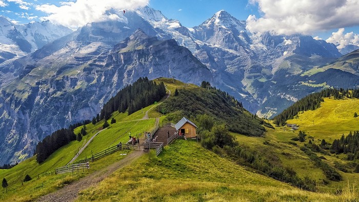 Wooden cabin on a grassy mountain ridge with snowy peaks