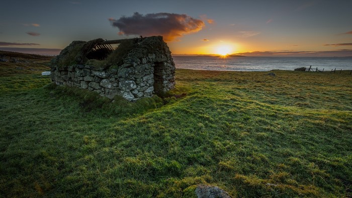 Ruined stone house on a grassy hill overlooking the sea at sunrise