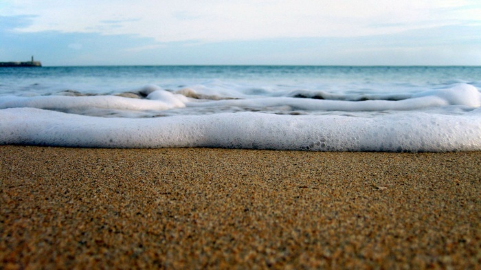 Ocean waves washing onto a sandy beach with a lighthouse in the distance