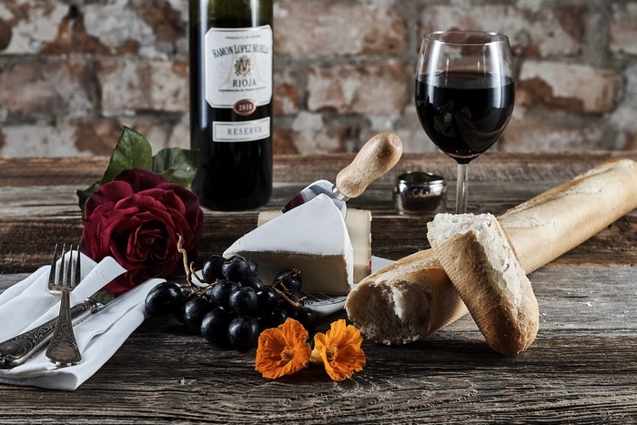 Wine, cheese, bread, grapes, rose, and cutlery on a wooden surface