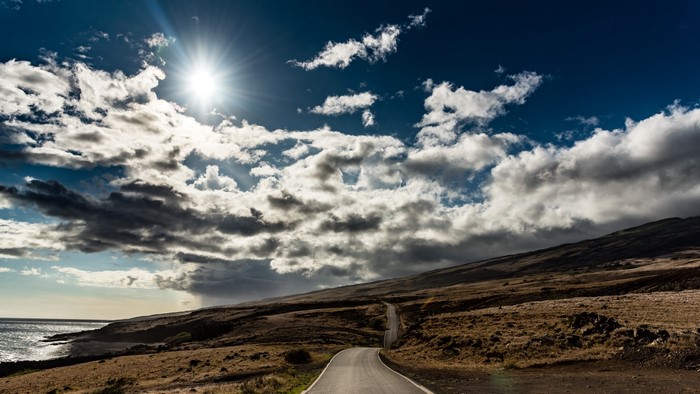 Road through a dry landscape under a cloudy sky with sun
