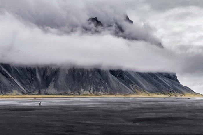 Person walking on a black sand beach with mountains and clouds