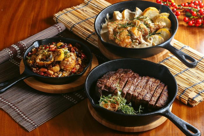 Three cast iron pans with steak, chicken, and seafood dishes