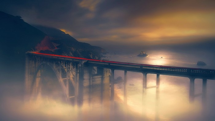 Bridge with car light trails over foggy ocean