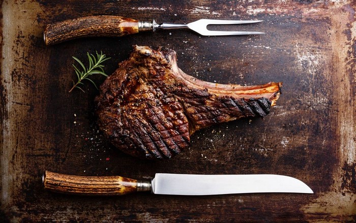 Tomahawk steak with rosemary, fork, and knife on a cutting board
