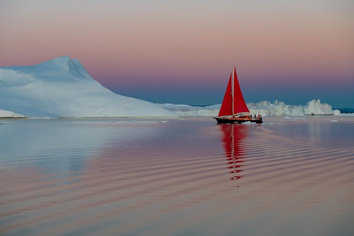 Sailboat with red sail on water near icebergs